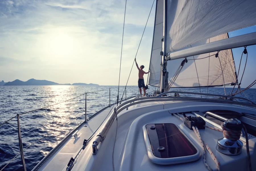 A man sailing a large sailboat off the shore of a mountainous coast.