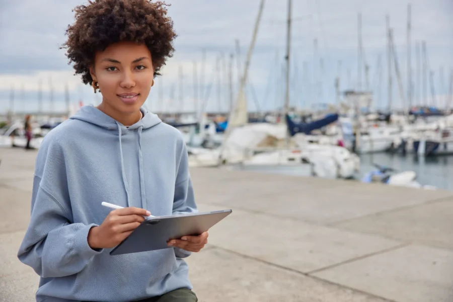 A person in a light blue hoodie holds a tablet and stylus, seated outdoors near a marina with sailboats in the background.