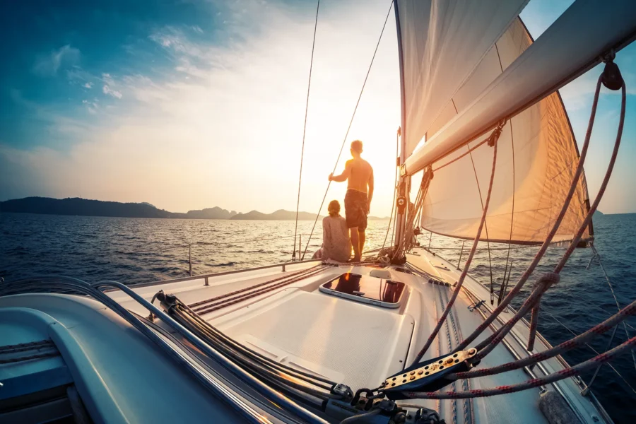 A man and woman stand on the deck of a sailboat at sunset, looking out over the ocean with the sails raised and distant land visible on the horizon.