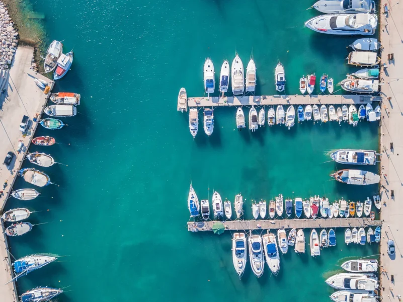 Aerial view of a marina with numerous boats and yachts docked along two parallel piers in clear turquoise water.