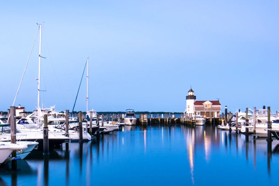 A marina with multiple docked boats and yachts in calm blue water, with a lighthouse and house in the background under a clear sky.