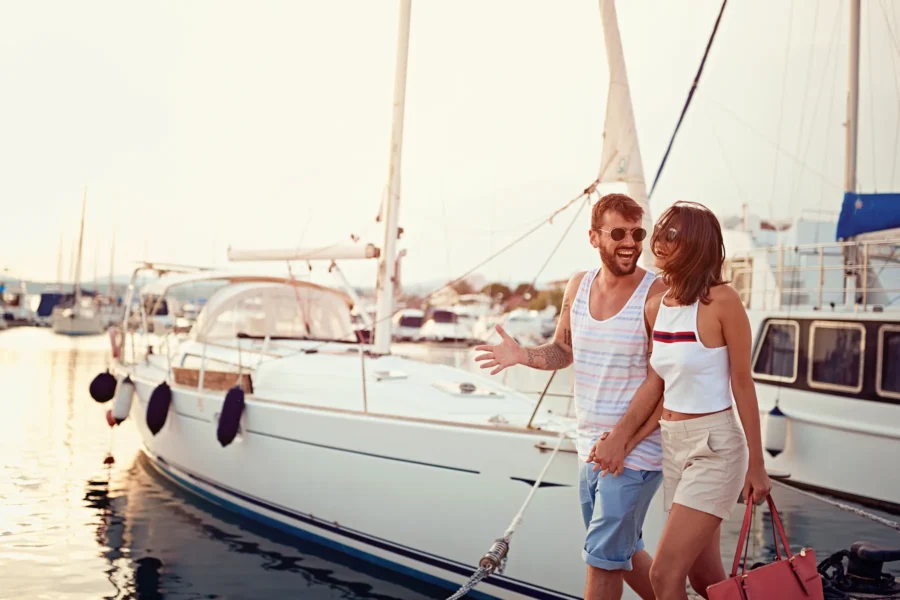A man and woman walk hand in hand on a marina dock, smiling and talking, with sailboats moored in the background.