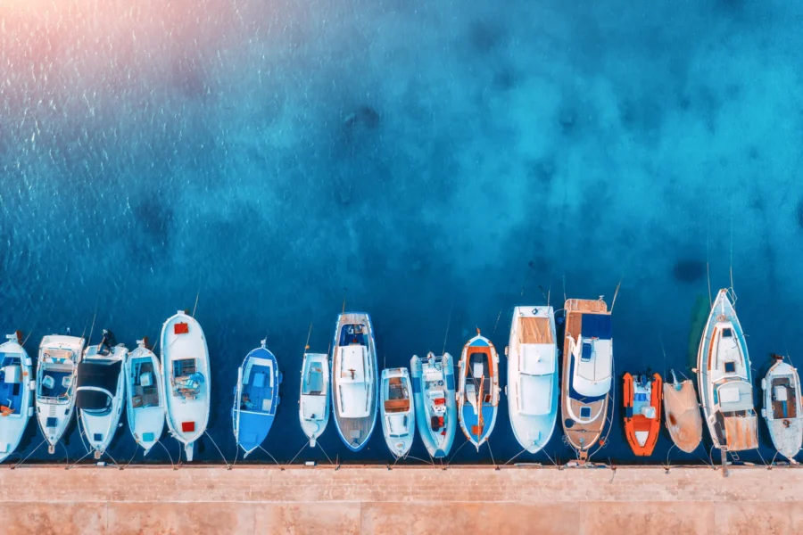 A row of various small boats is docked along a concrete pier in clear blue water, viewed from above.