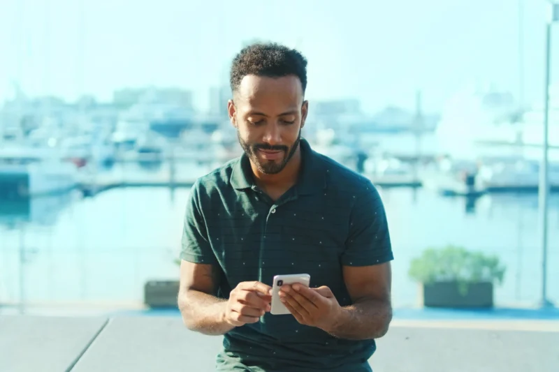 Man wearing a dark green polo shirt sits outdoors by a marina, looking at and using a smartphone with boats visible in the background.