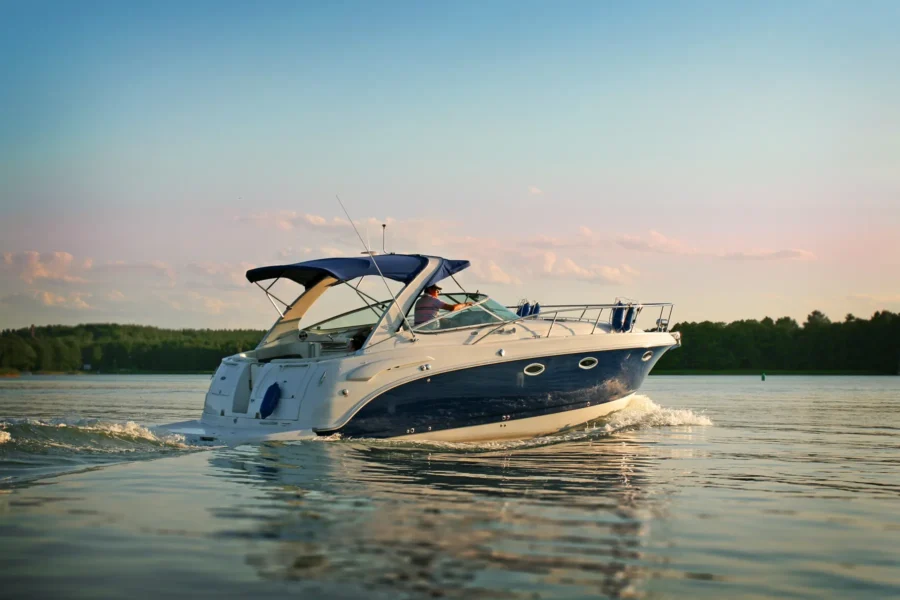 A white and blue motorboat with a canopy moves across calm water near a forested shoreline under a clear sky at sunset.
