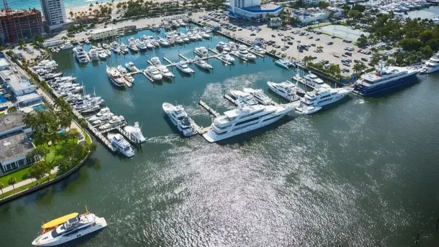 Aerial view of a marina with numerous docked yachts and boats, adjacent to a parking lot and buildings near the waterfront.